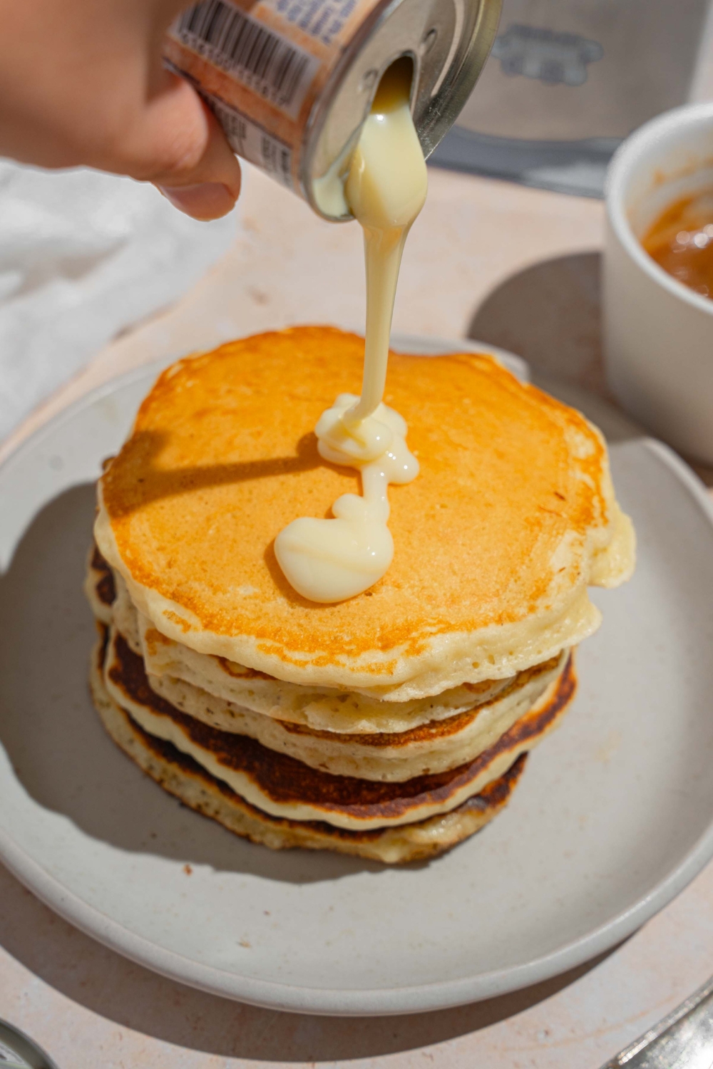 A stack of pancakes on a white plate with a can of condensed milk being poured onto the pancakes. The plate is on a tan counter with a small bowl of caramel sauce.