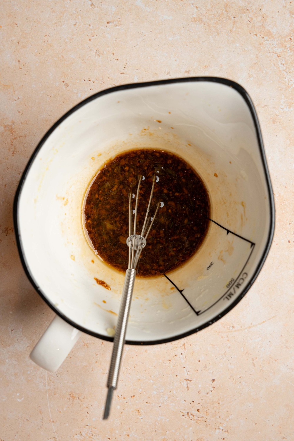 A ceramic pitcher with a whisk mixing ingredients to make carrot salad dressing including soy sauce, rice vinegar, sesame oil, garlic, and more. The pitcher is on a tan counter.