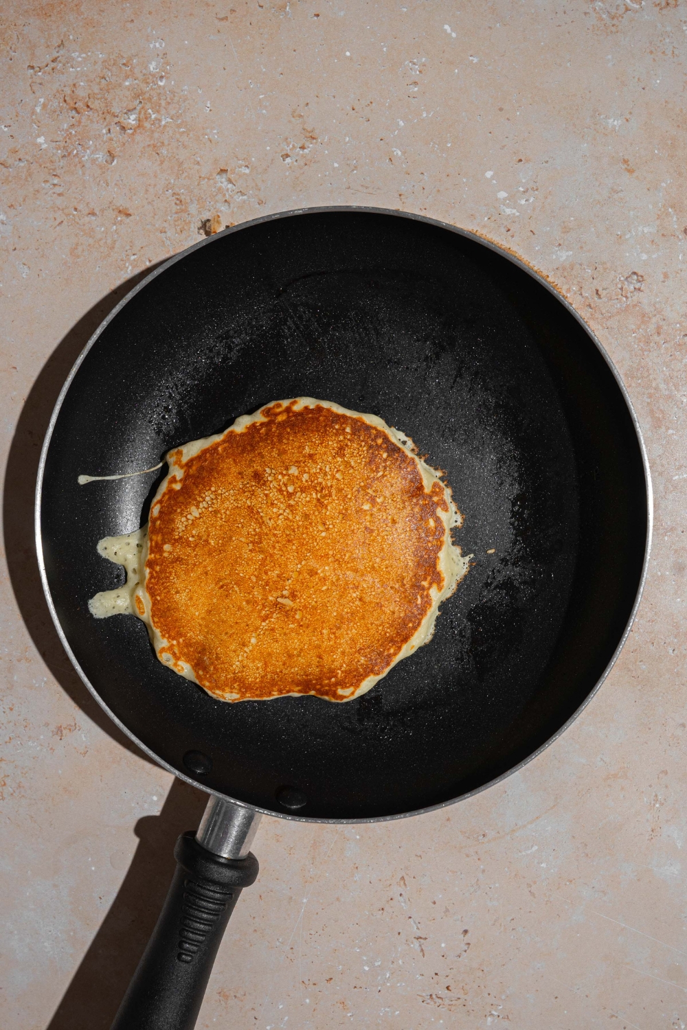 A skillet with a flipped cooked pancake. The skillet is on a tan counter.