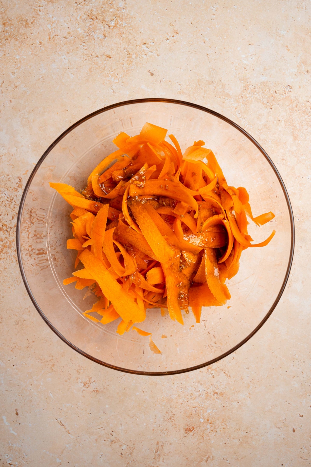 A glass bowl with shredded carrot ribbons tossed in dressing. The bowl is on a tan counter.