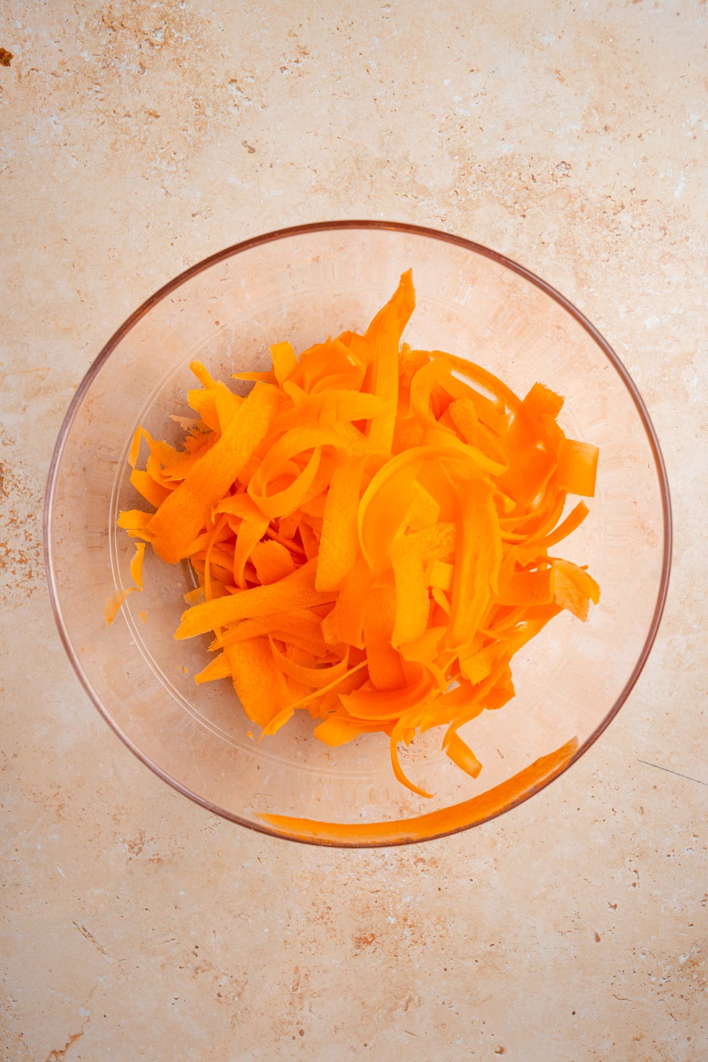 A glass bowl with shredded carrot ribbons. The bowl is on a tan counter.