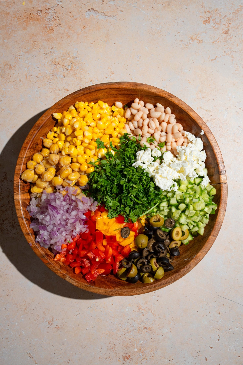 A wooden bowl with ingredients to make Mediterranean dense bean salad including navy beans, chickpeas, corn, red onion, bell peppers, cucumber, feta cheese, and fresh parsley. The bowl is on a tan counter.