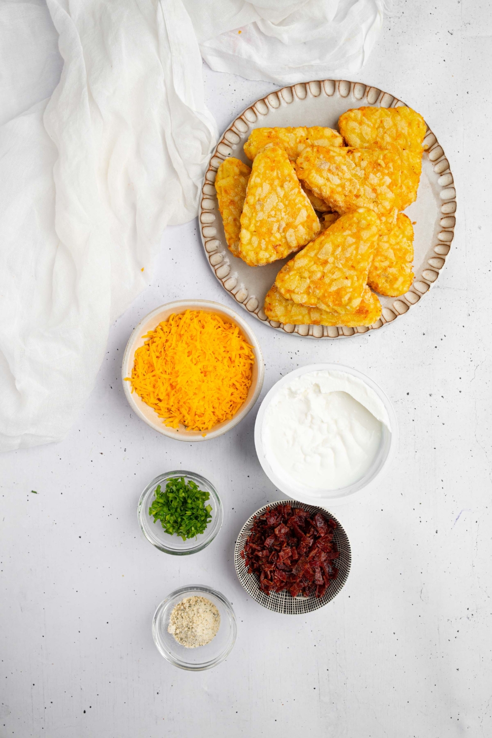 An overhead shot of several bowls in various sizes containing ingredients to make loaded hashbrowns including hashbrown patties, shredded cheddar cheese, sour cream, ranch seasoning, crumbled bacon, and chives.