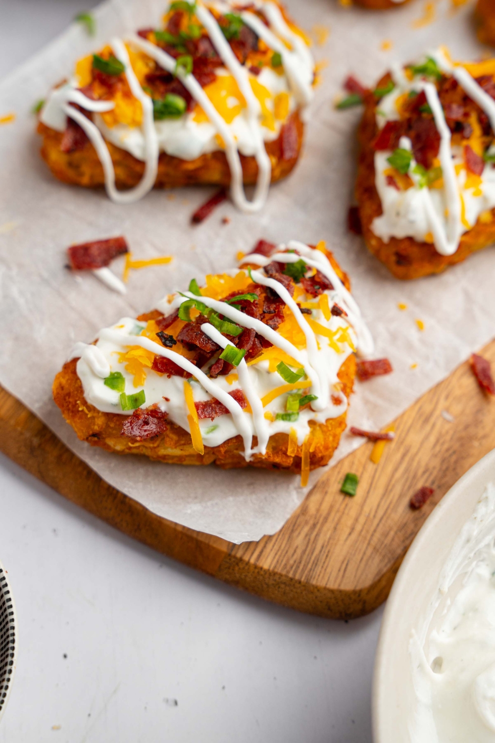 Several loaded hashbrowns topped with sour cream mixture, cheddar cheese, and crumbled bacon on a wooden board lined with parchment paper. The hashbrowns are drizzled with sour cream and garnished with sliced chives.