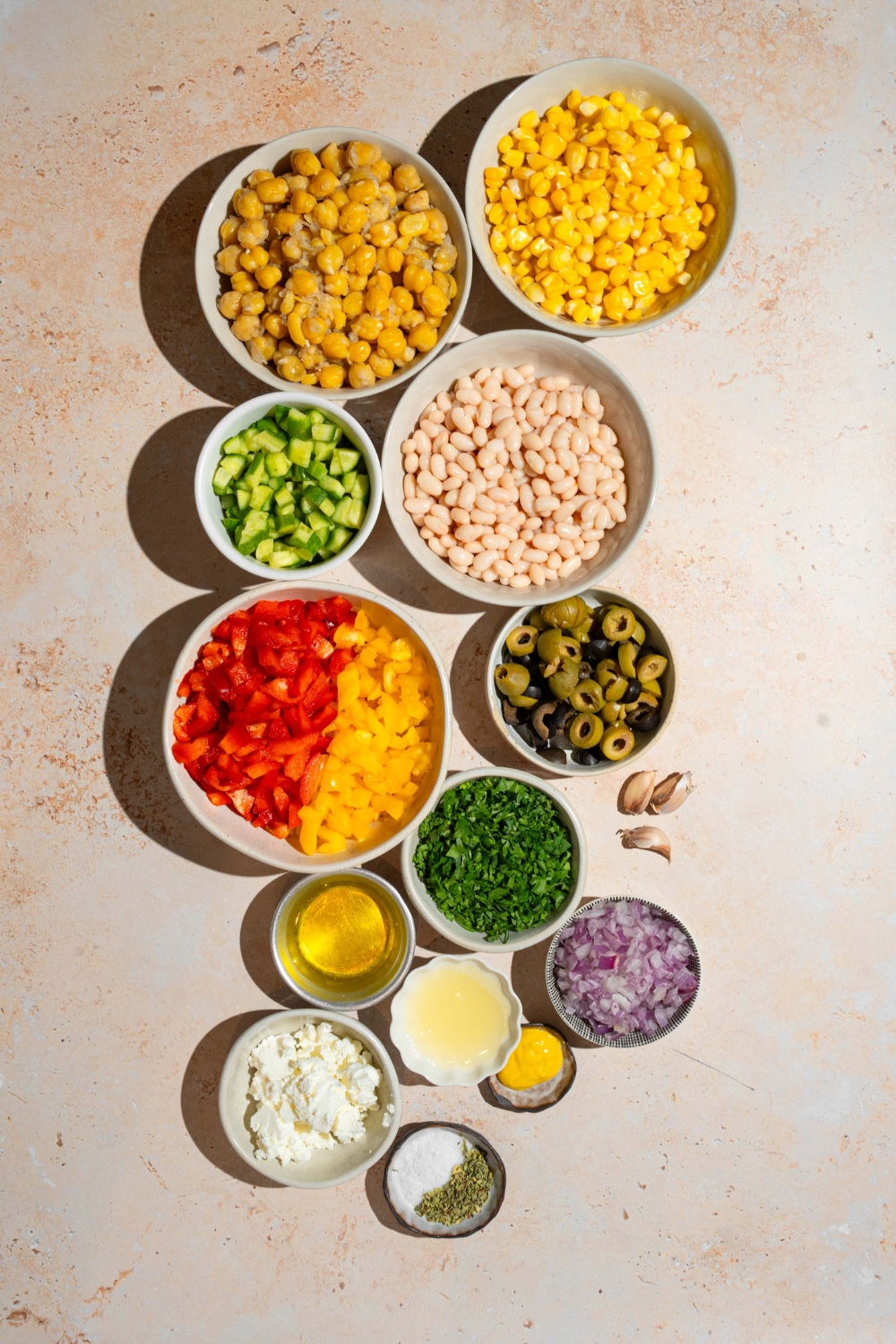 An overhead shot of several bowls in various sizes containing ingredients to make Mediterranean dense bean salad including chickpeas, navy beans, corn, cucumber, bell peppers, olvies, red onion, fresh parsley, and more.