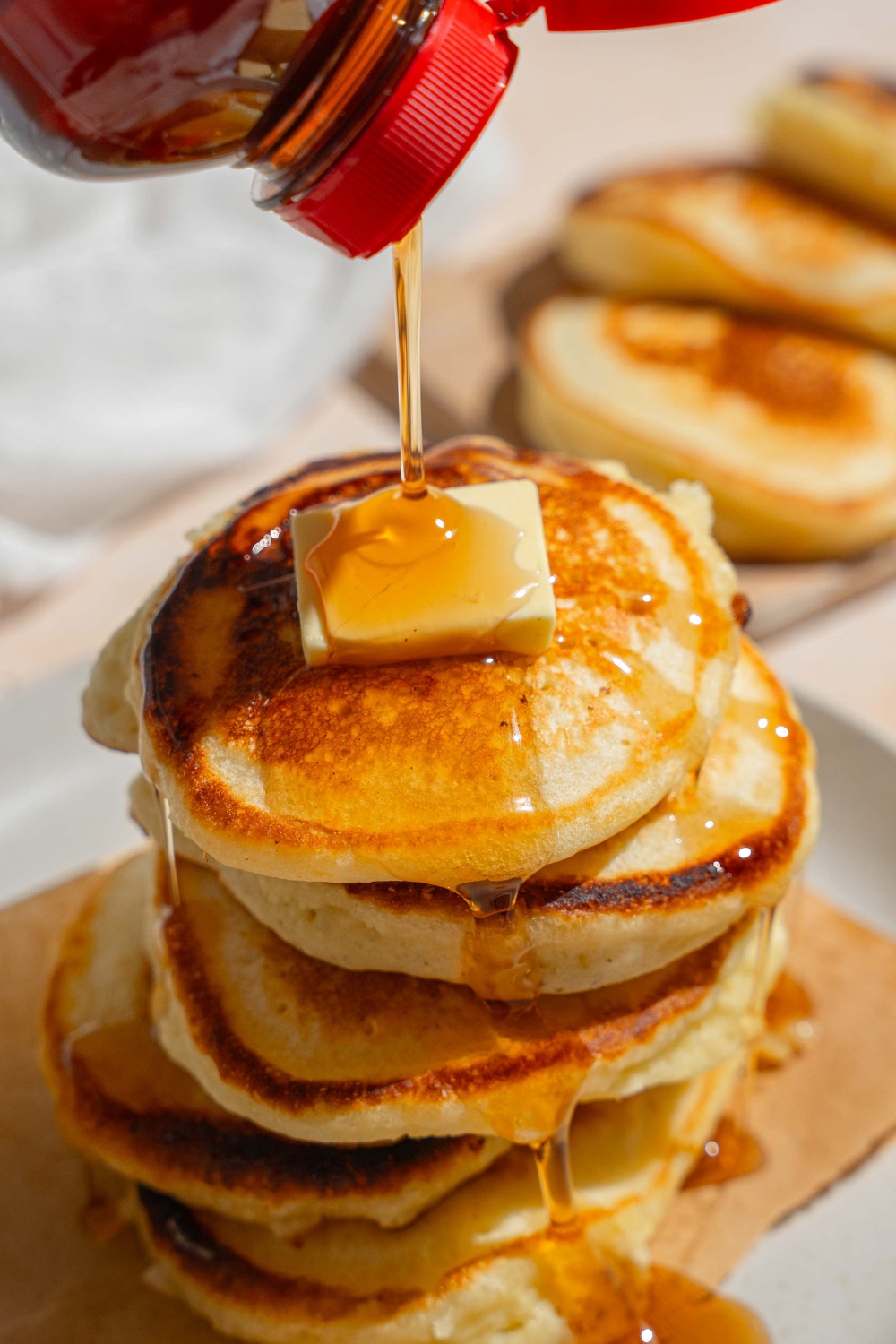 A white plate lined with a paper napkin with a stack of crispy pancakes topped with a pad of butter. A bottle is pouring syrup over the pancakes. The plate is on a tan counter with a board of pancakes and white cloth napkin.