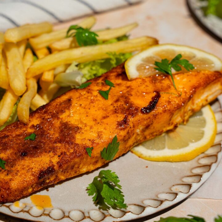 A ceramic plate with frozen air fryer salmon served with lettuce and french fries and garnished with fresh parsley and lemon. The plate is on a tan counter with a white striped napkin.