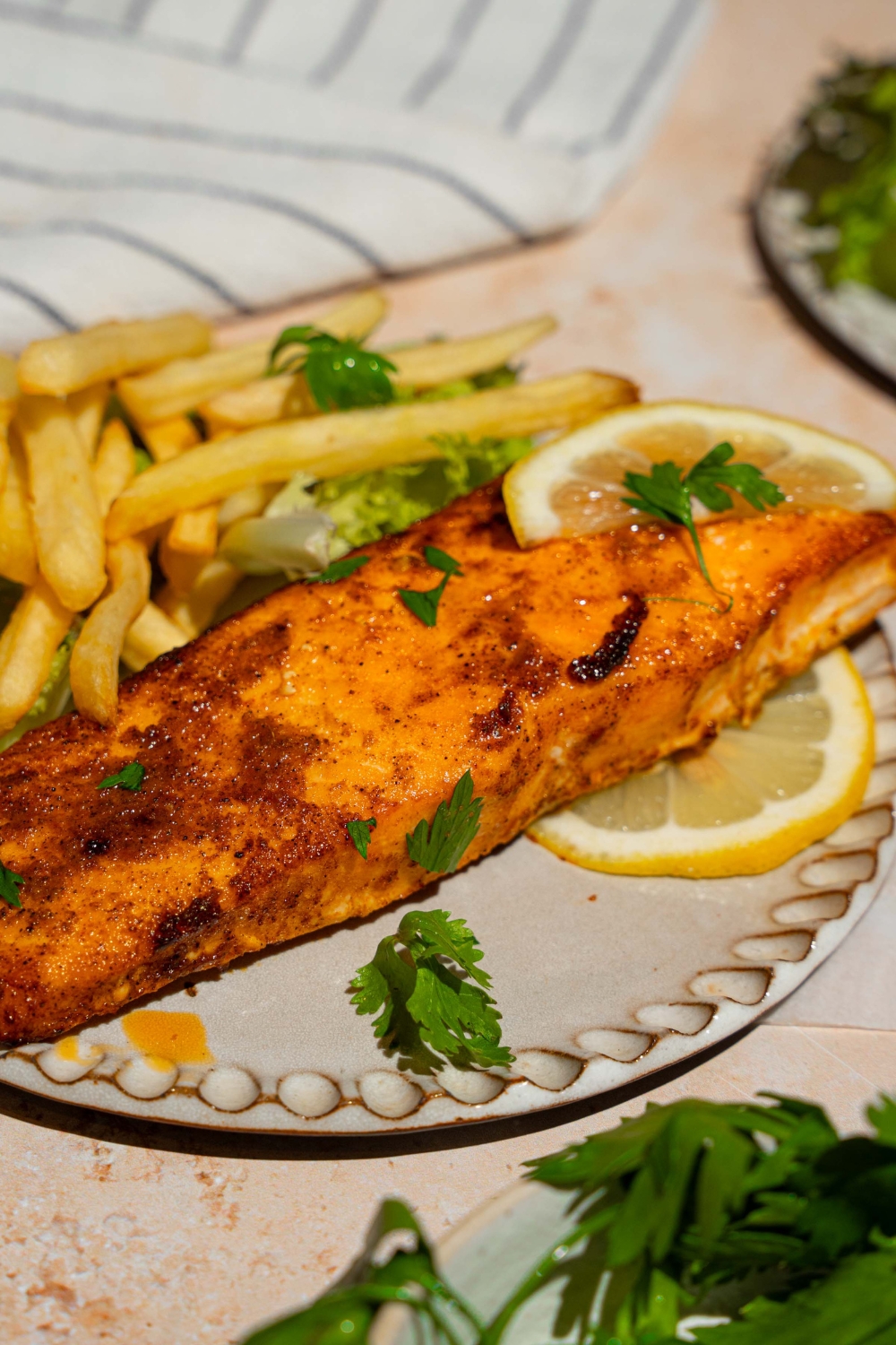 A ceramic plate with frozen air fryer salmon served with lettuce and french fries and garnished with fresh parsley and lemon. The plate is on a tan counter with a white striped napkin.