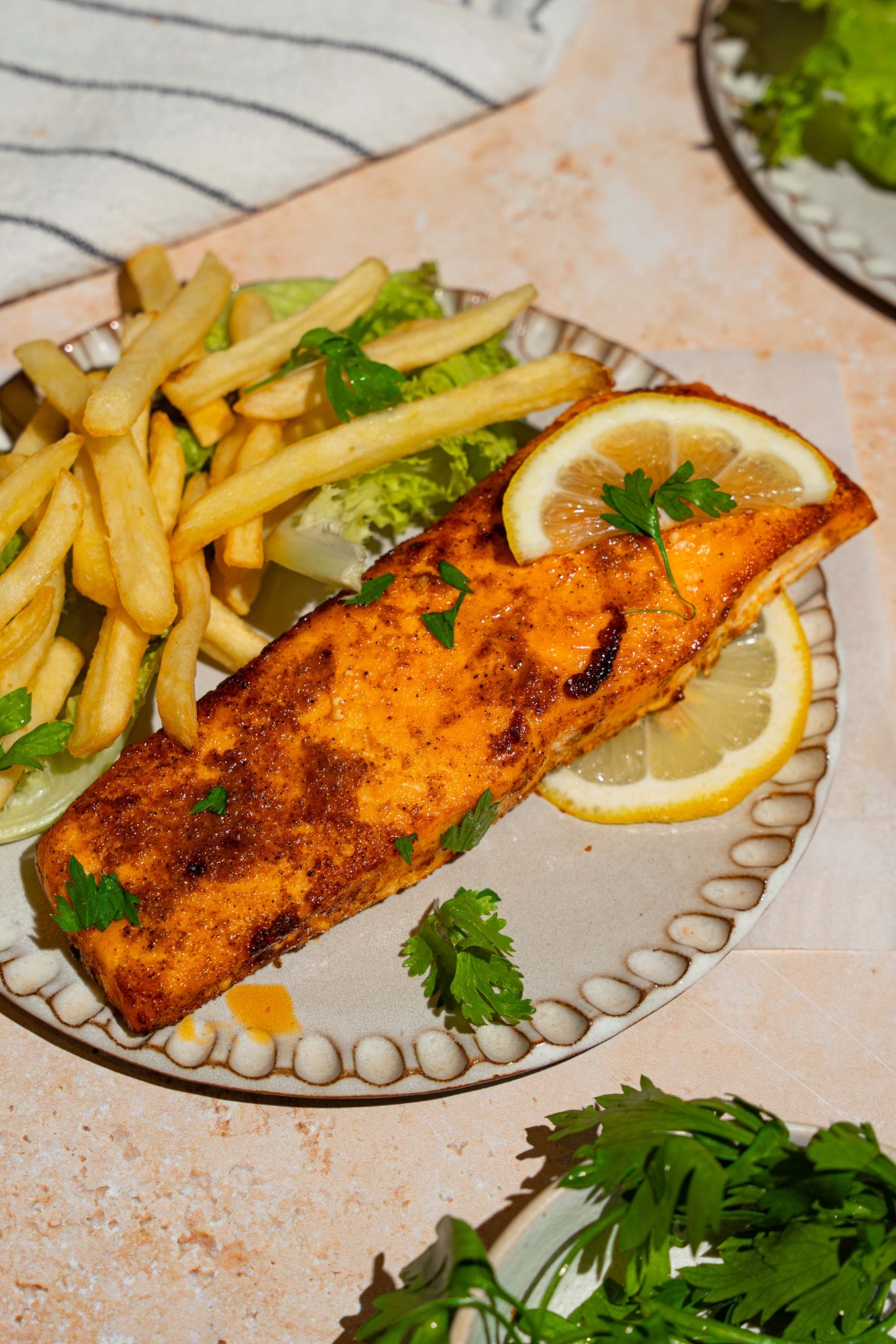 A ceramic plate with frozen air fryer salmon served with lettuce and french fries and garnished with fresh parsley and lemon. The plate is on a tan counter with a white striped napkin.