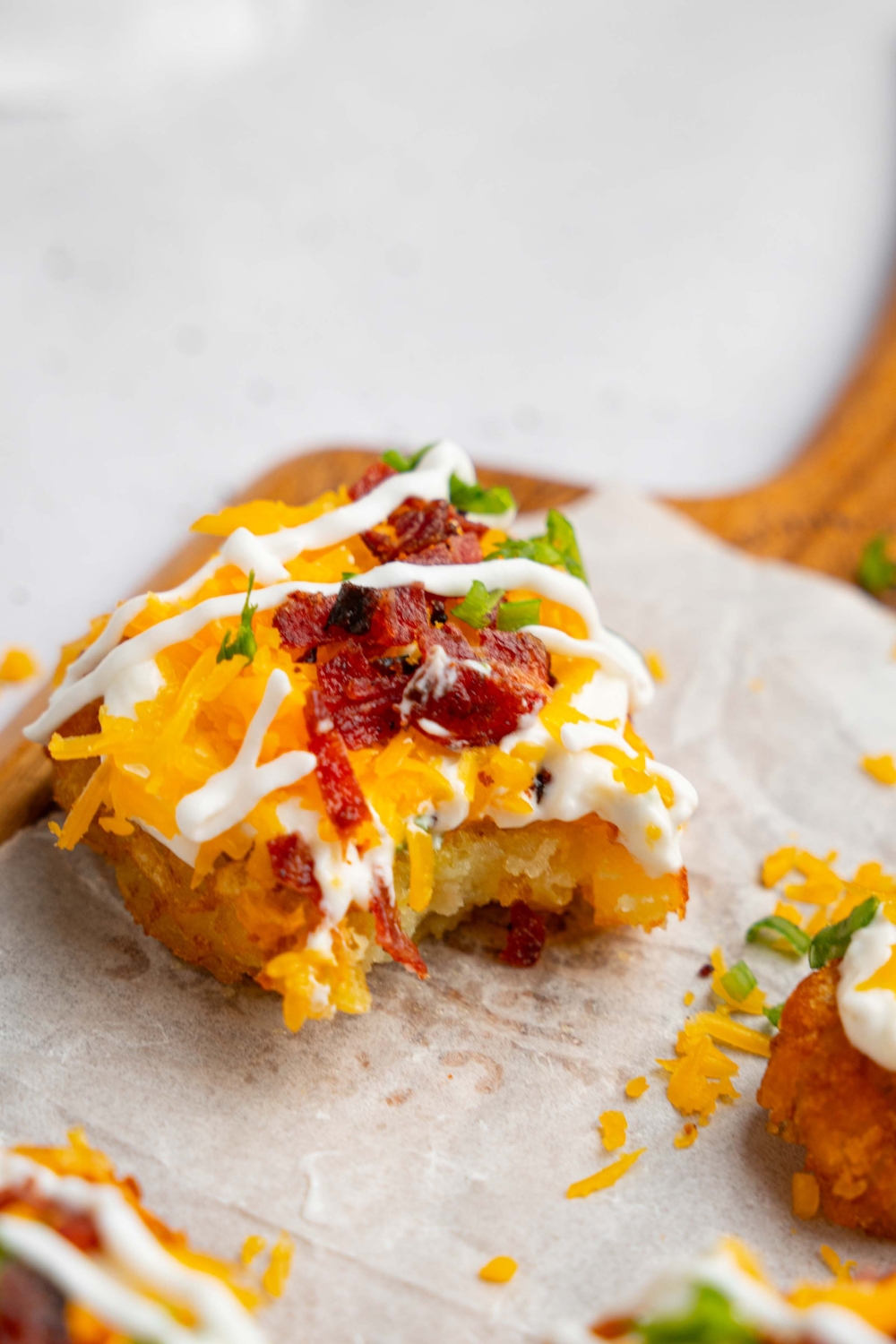Several loaded hashbrowns topped with sour cream mixture, cheddar cheese, and crumbled bacon on a wooden board lined with parchment paper. The hashbrowns are drizzled with sour cream and garnished with sliced chives. A bite is taken from one of the hashbrowns.
