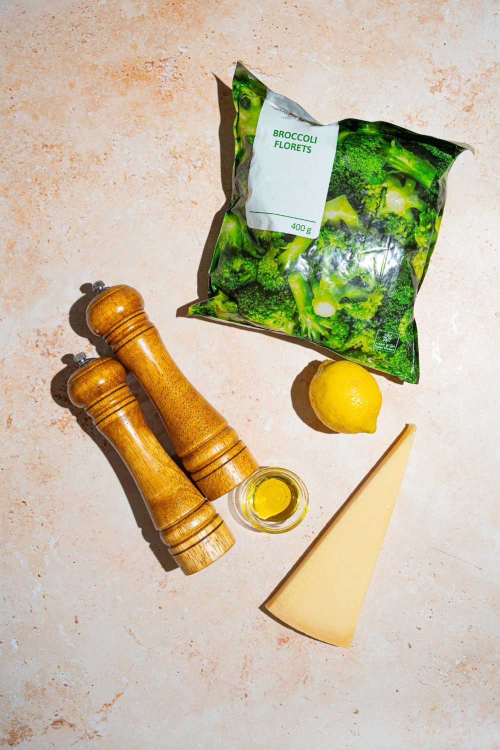 An overhead shot of ingredients to make roasted frozen broccoli including roasted broccoli, lemon, olive oil, a wedge of parmesan cheese, and a salt and pepper mill.