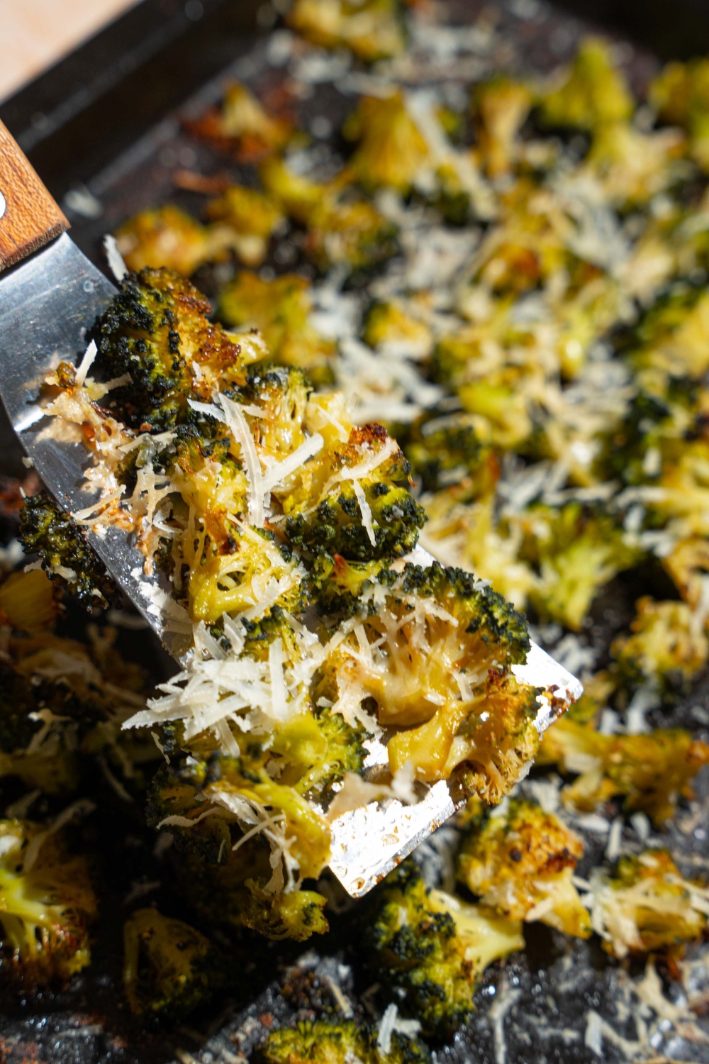 A roasting pan with roasted frozen broccoli with grated parmesan. A spatula is taking a portion of broccoli.