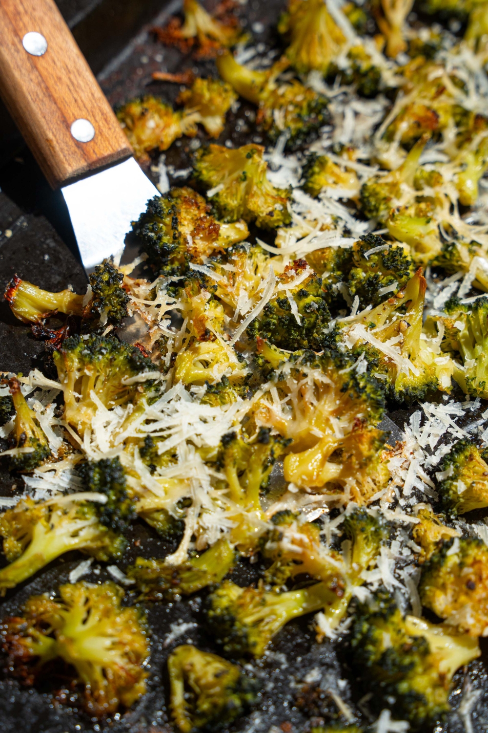A roasting pan with roasted frozen broccoli with grated parmesan. A spatula is taking a portion of broccoli.