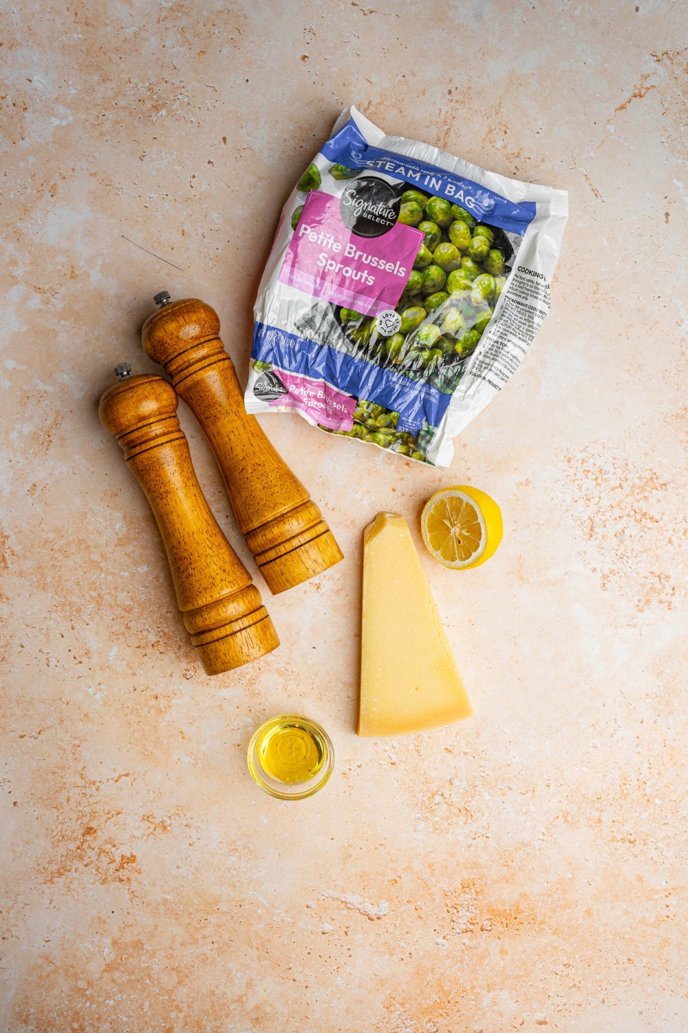 An overhead shot of ingredients to make roasted brussel sprouts including frozen brussel sprouts, lemon, parmesan cheesse wedge, olive oil, and a salt and pepper mill.