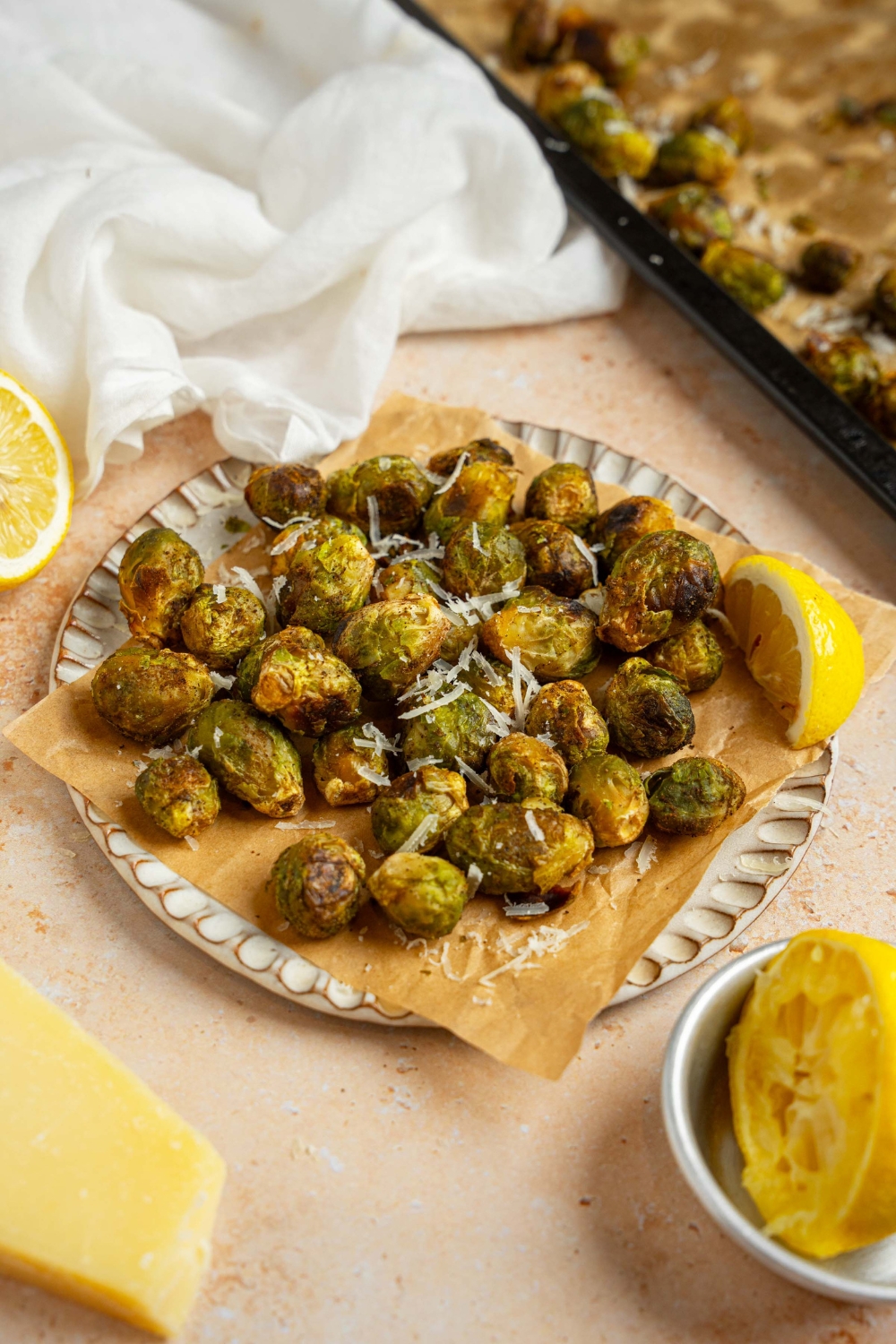 A ceramic plate lined with parchment paper garnished with grated parmesan. The plate is on a tan counter with a tray of roasted brussel sprouts and white cloth napkin.