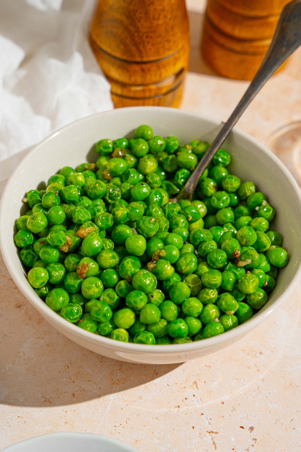 A white bowl with cooked frozen peas tossed in seasonings. There is a spoon in the bowl. The bowl is on a tan counter with a white cloth napkin.