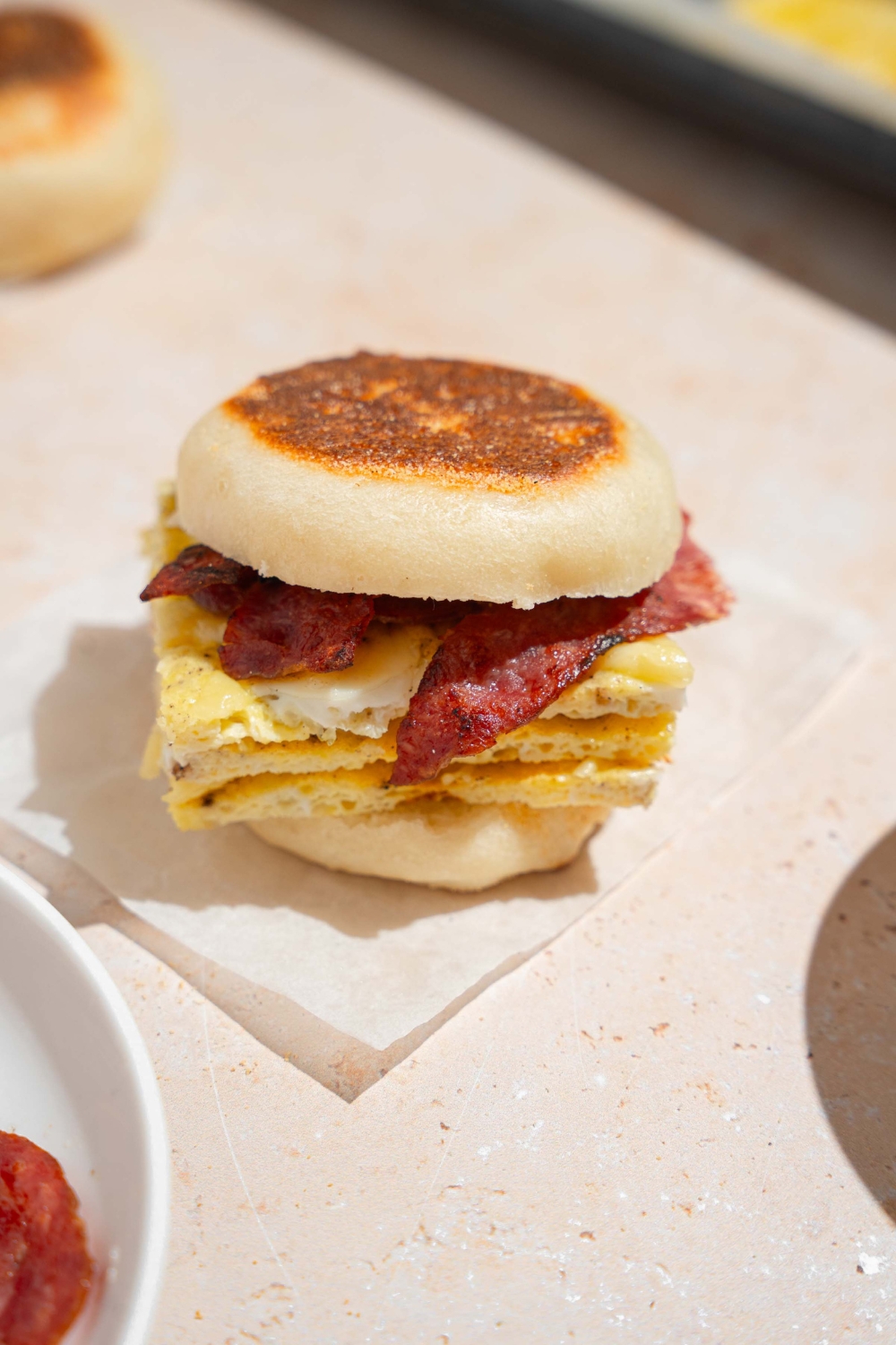 A sheet pan egg sandwich with eggs, bacon strips, and cheese in between an english muffin. The sandwich is on a piece of parchment paper on a tan counter.