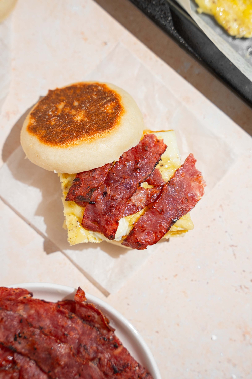 An open sheet pan egg sandwich with bacon strips, cheese, on an english muffin. The sandwich is on a piece of parchment paper on a tan counter with a bowl of bacon strips.