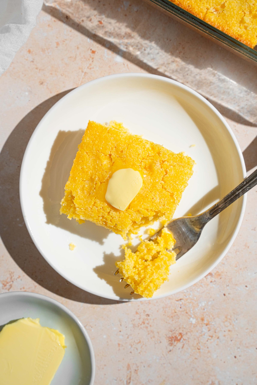 A slice of Fat Daddy's cornbread topped with a pat of butter. There is a fork taking a bite of bread. The slice is on a white plate on a tan with a baking dish of cornbread.