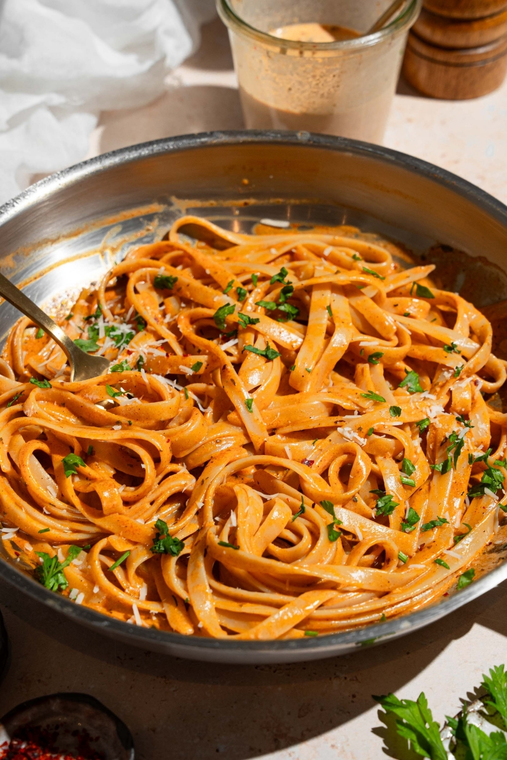 A skillet with fettuccine pasta tossed in cajun cream sauce. The pasta is garnished with fresh parsley and grated cheese. There is a fork in the pasta. The skillet is on a tan counter with a jar of cream sauce.