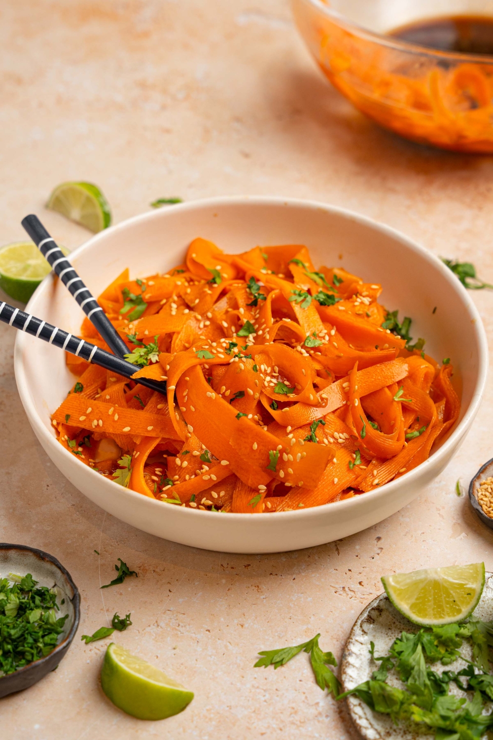 A bowl of carrot salad garnished with sesame seeds and fresh cilantro. There is a pair of chopsticks in the bowl. The bowl is on a tan counter with small plates of garnishes and lime wedges.