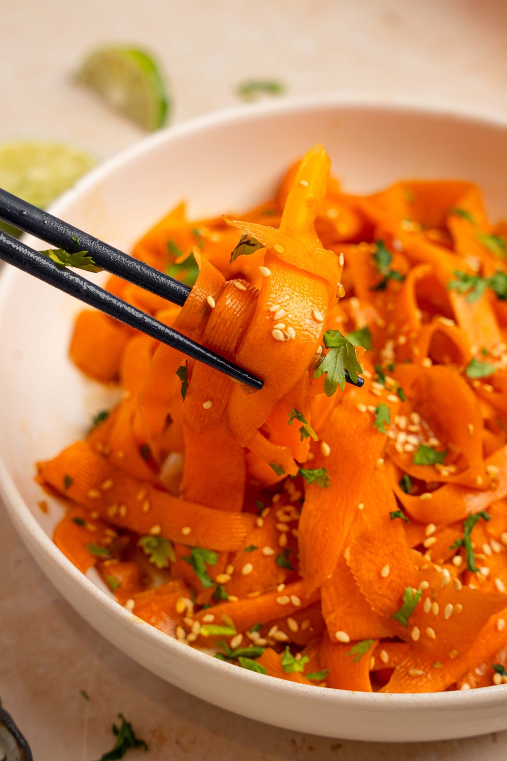 A bowl of carrot salad garnished with sesame seeds and fresh cilantro. There is a pair of chopsticks taking a bite from the bowl. The bowl is on a tan counter with small plates of garnishes and lime wedges.