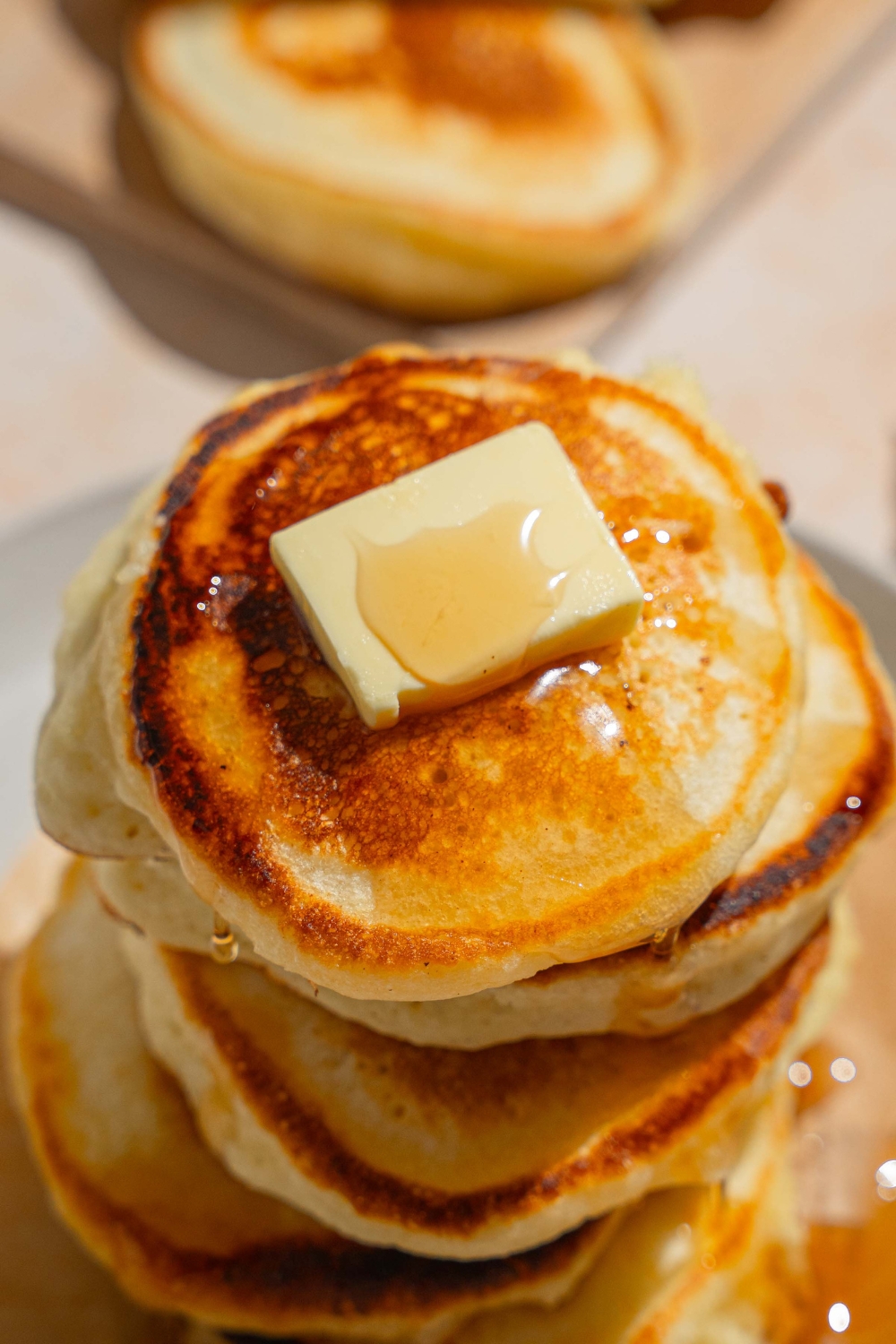 A stack of crispy pancakes on a white plate lined with a napkin. The pancakes are drizzled with syrup and topped with a pad of butter. The plate is on a tan counter with a board of pancakes.