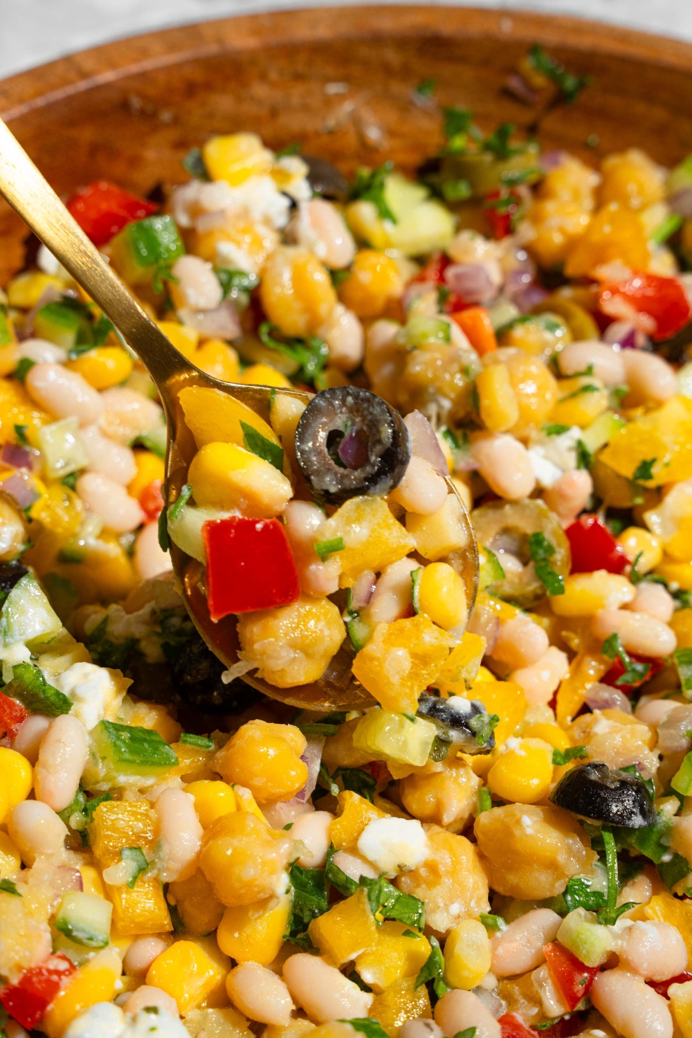 A wooden bowl with Mediterranean dense bean salad including black olives, corn, navy beans, chickpeas, bell peppers, feta cheese, and cucumber garnished with fresh parsley. There is a spoon with a bite of salad.