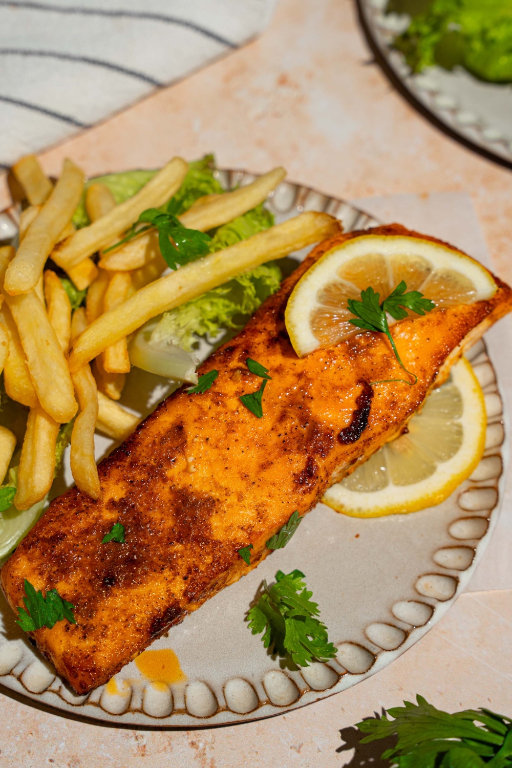 A ceramic plate with frozen air fryer salmon served with lettuce and french fries and garnished with fresh parsley and lemon. The plate is on a tan counter with a white striped napkin.