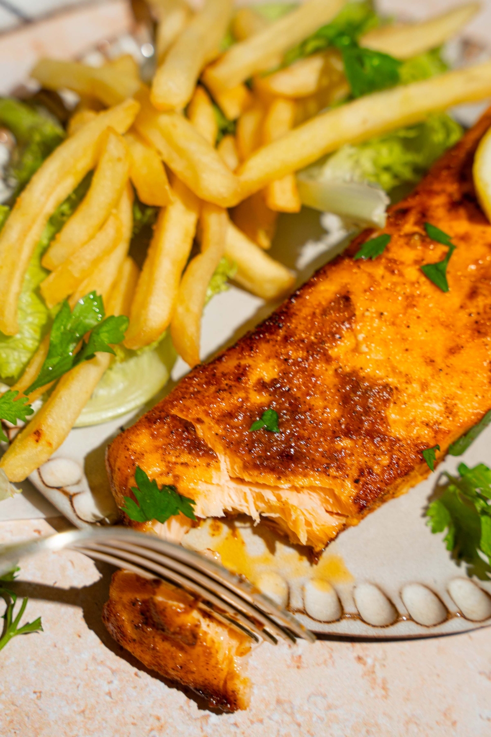 A ceramic plate with frozen air fryer salmon served with lettuce and french fries and garnished with fresh parsley and lemon. A fork is taking a bite of salmon.