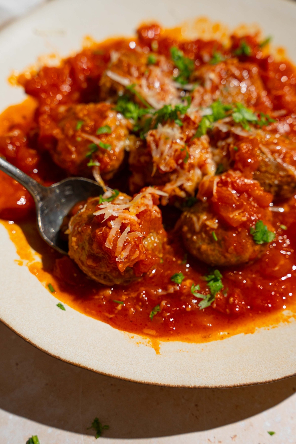 A white plate with frozen meatballs cooked in tomato sauce topped with grated cheese and fresh parsley. A spoon is scooping a meatball from the plate.