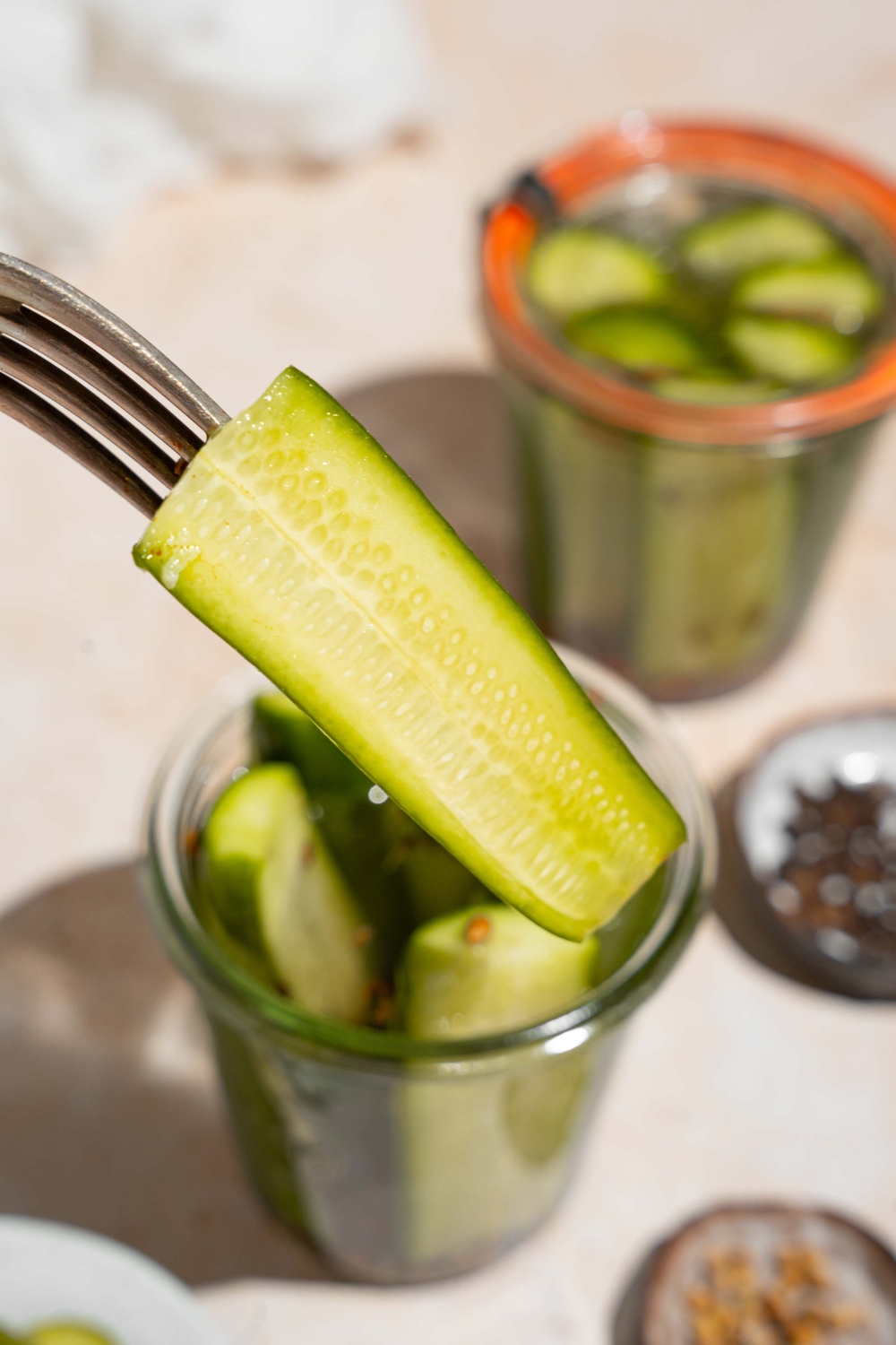 A close up of a fork with a homemade Claussen pickle with a jar of pickles blurred in the background. The jar is on a tan counter with an additional jar and plates of seasonings.