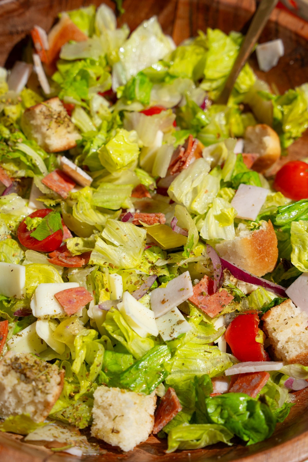 A wooden salad bowl of copycat Jersey Mike's sub in a tub including mixed lettuce, tomatoes, provolone cheese, diced meats, and bread croutons. There is a fork in the salad.