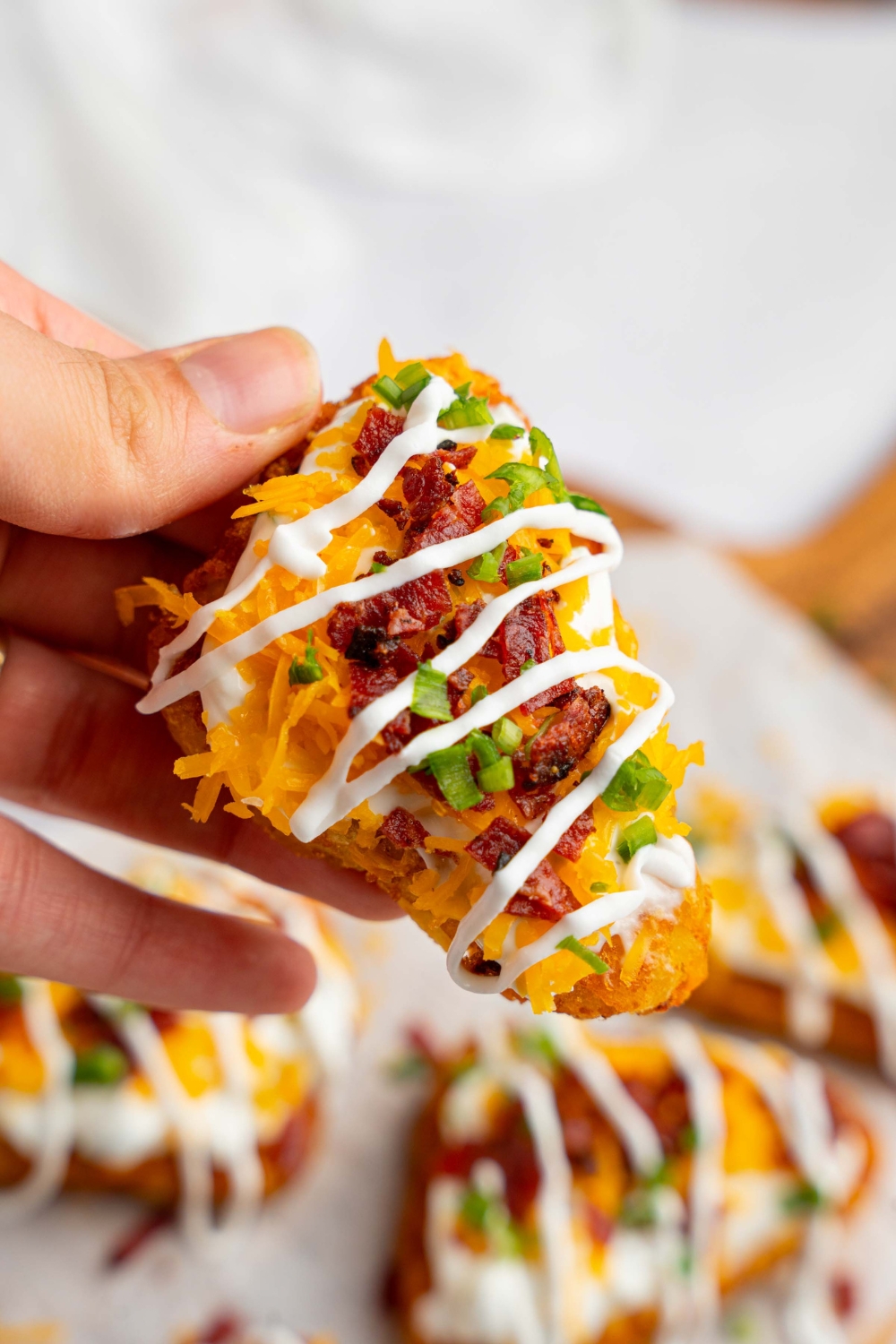 A close up of a hand holding a loaded hashbrown drizzled with sour cream and sliced chives. There is a board of hashbrowns blurred in the background.