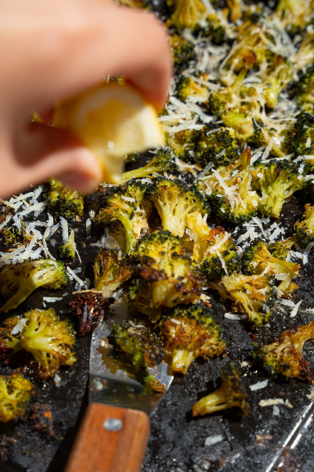 A roasting pan with roasted frozen broccoli with grated parmesan and a spatula. There is a hand squeezing lemon over the broccoli.