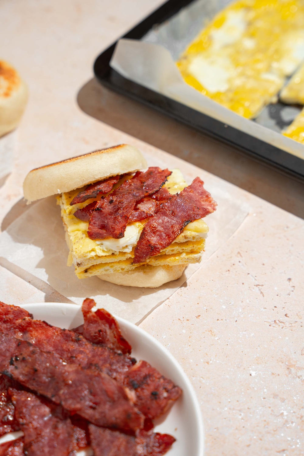 An open sheet pan egg sandwich with bacon strips, cheese, on an english muffin. The sandwich is on a piece of parchment paper on a tan counter with a bowl of bacon strips.