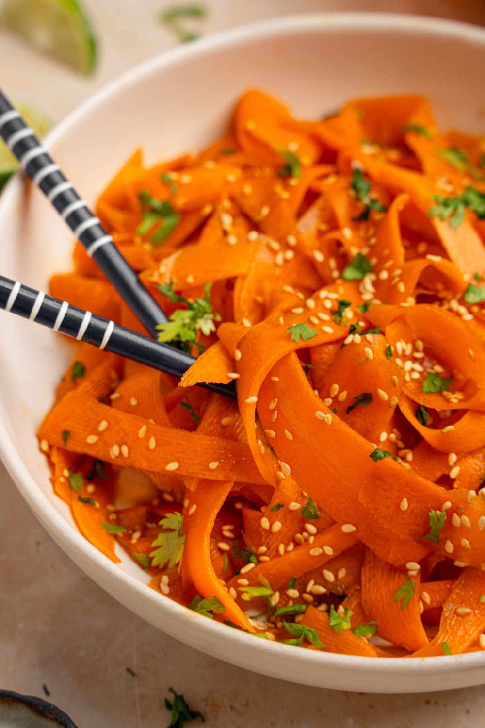A bowl of carrot salad garnished with sesame seeds and fresh cilantro. There is a pair of chopsticks in the bowl. The bowl is on a tan counter with small plates of garnishes and lime wedges.