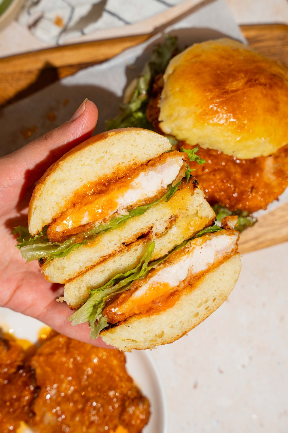 A hand holding a hot honey chicken sandwich sliced in half. There is a wooden board with an additional sandwich in the background.