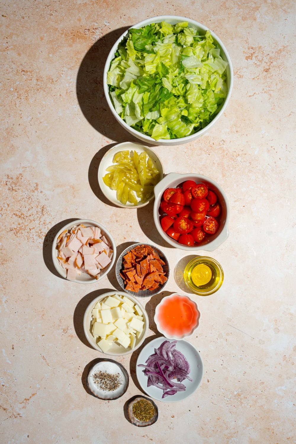An overhead shot of several bowls containing ingredients to make copycat Jersey Mike's sub in a tub including chopped lettuce, tomatoes, diced meats, red onion, provolone cheese, oil, and seasonings.