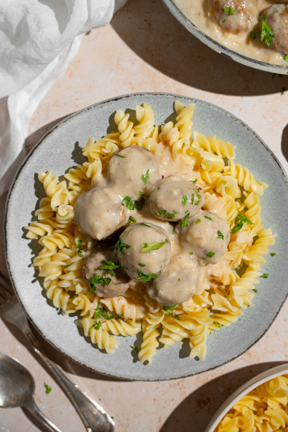 A ceramic plate with pasta topped with Swedish meatballs garnished with fresh parsley. The plate is on a tan counter with a fork and white cloth napkin.