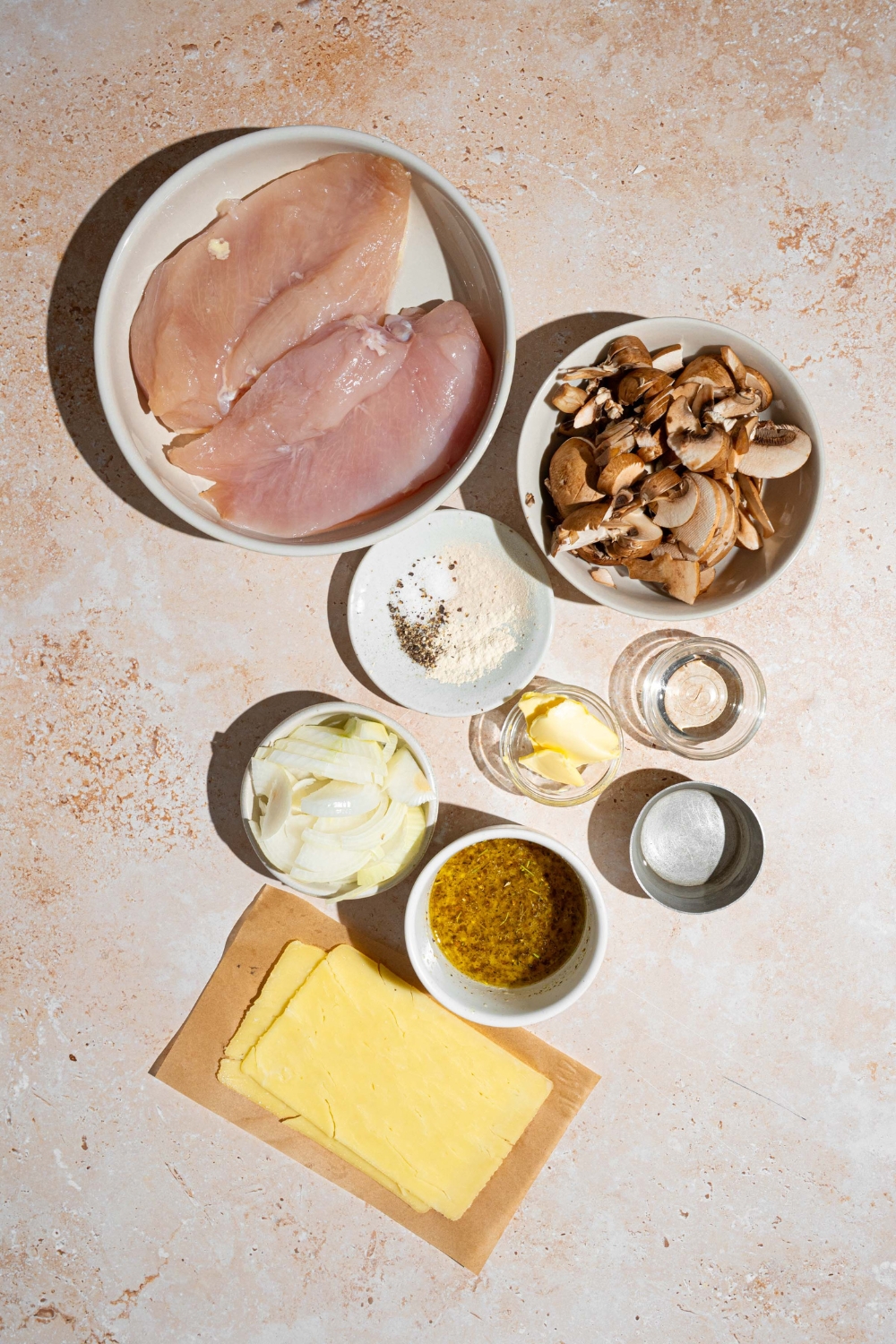 An overhead shot of several bowls in various sizes containing ingredients to make Texas Roadhouse smothered chicken including chicken breats, mushrooms, white onion, butter, monterey jack cheese, and seasonings.