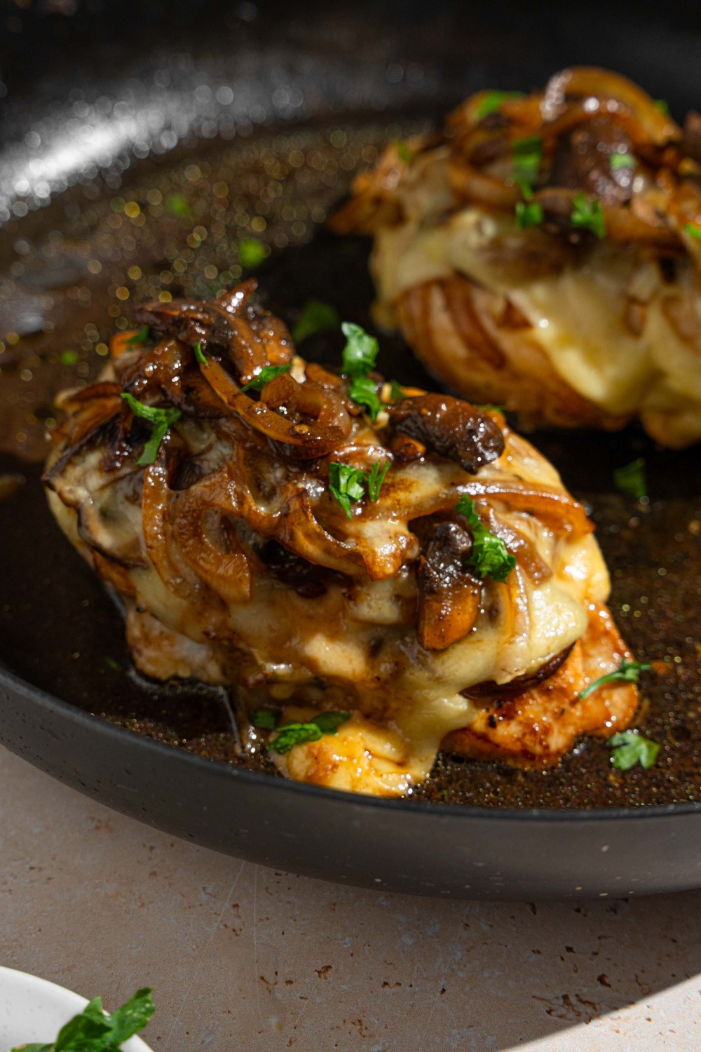 Two Texas Roadhouse smothered chicken breasts on a skillet in juices and garnished with fresh parsley. The chicken is topped with melted cheese and sautéed vegetables.