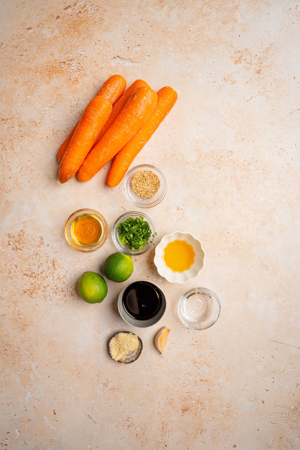 An overhead shot of several bowls in various sizes containing ingredients to make carrot salad including whole carrots, limes, soy sauce, rice vinegar, maple syrup, sesame oil, and more.