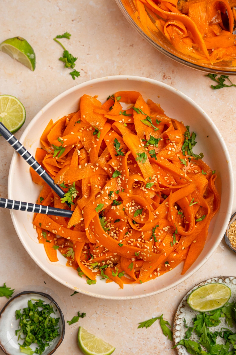 A bowl of carrot salad garnished with sesame seeds and fresh cilantro. There is a pair of chopsticks in the bowl. The bowl is on a tan counter with small plates of garnishes and lime wedges.