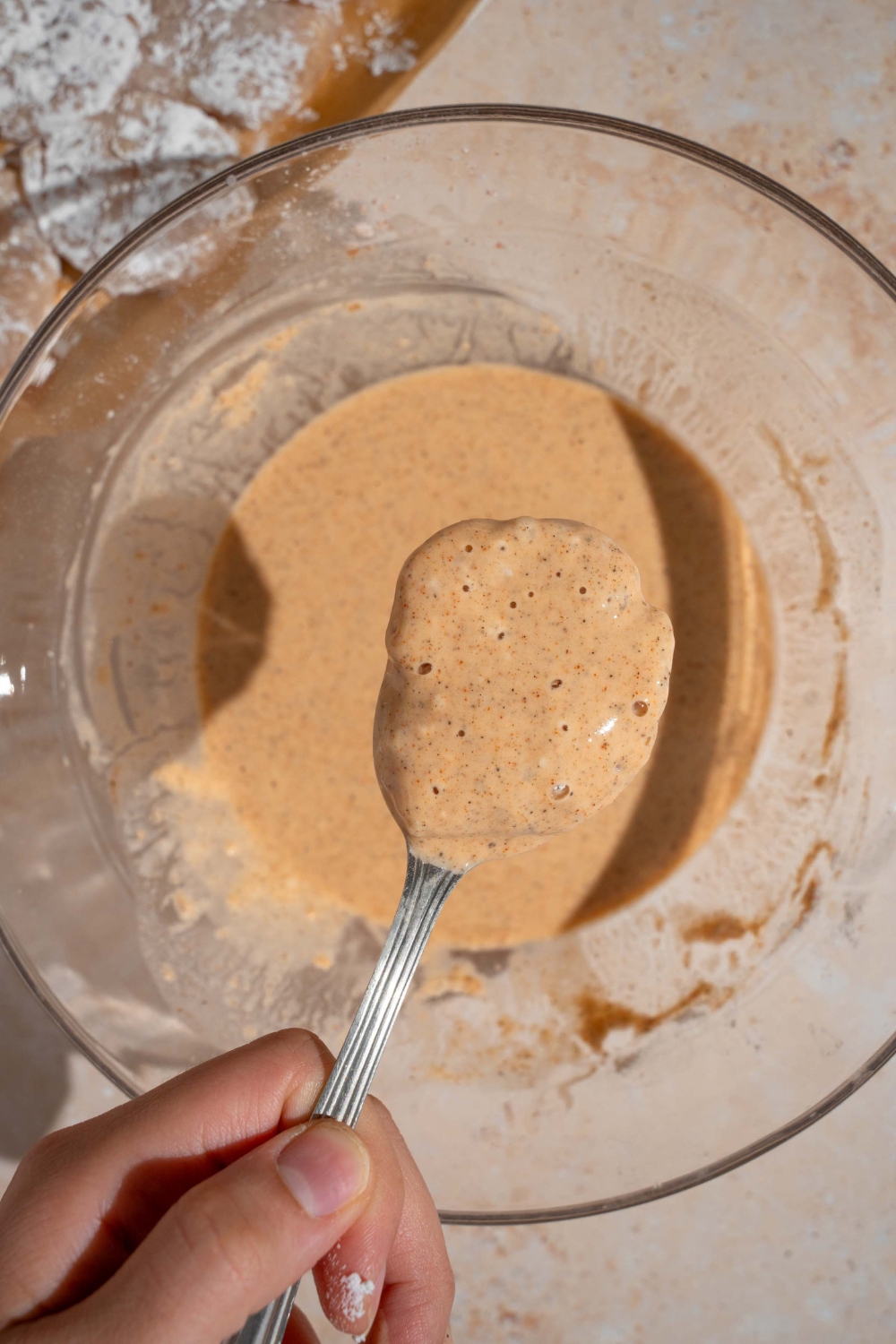 A spoon with a chicken nugget that is coated in liquid batter on it above a bowl of the batter.