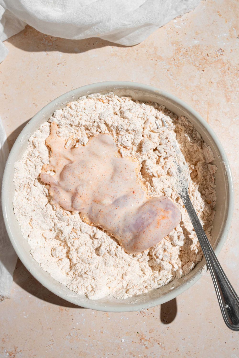 A raw chicken thigh in a white bowl that is filled with a flower mixture. The bowl is on top of the white counter.