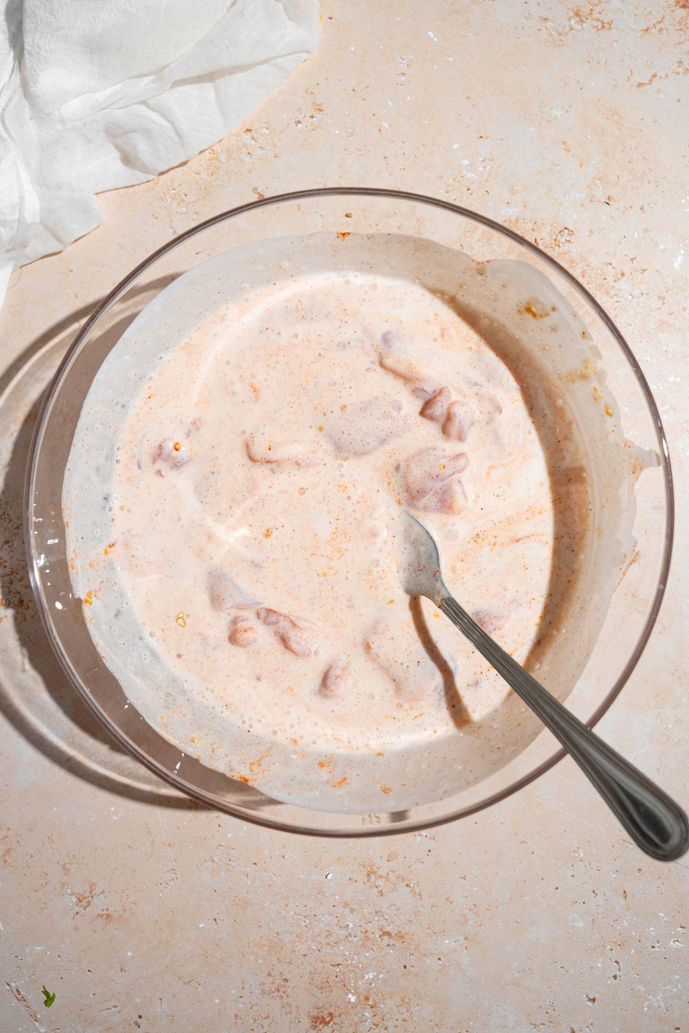 A spoon in a bowl of buttermilk marinade with chicken thighs in it. The bowl is on top of a white counter.