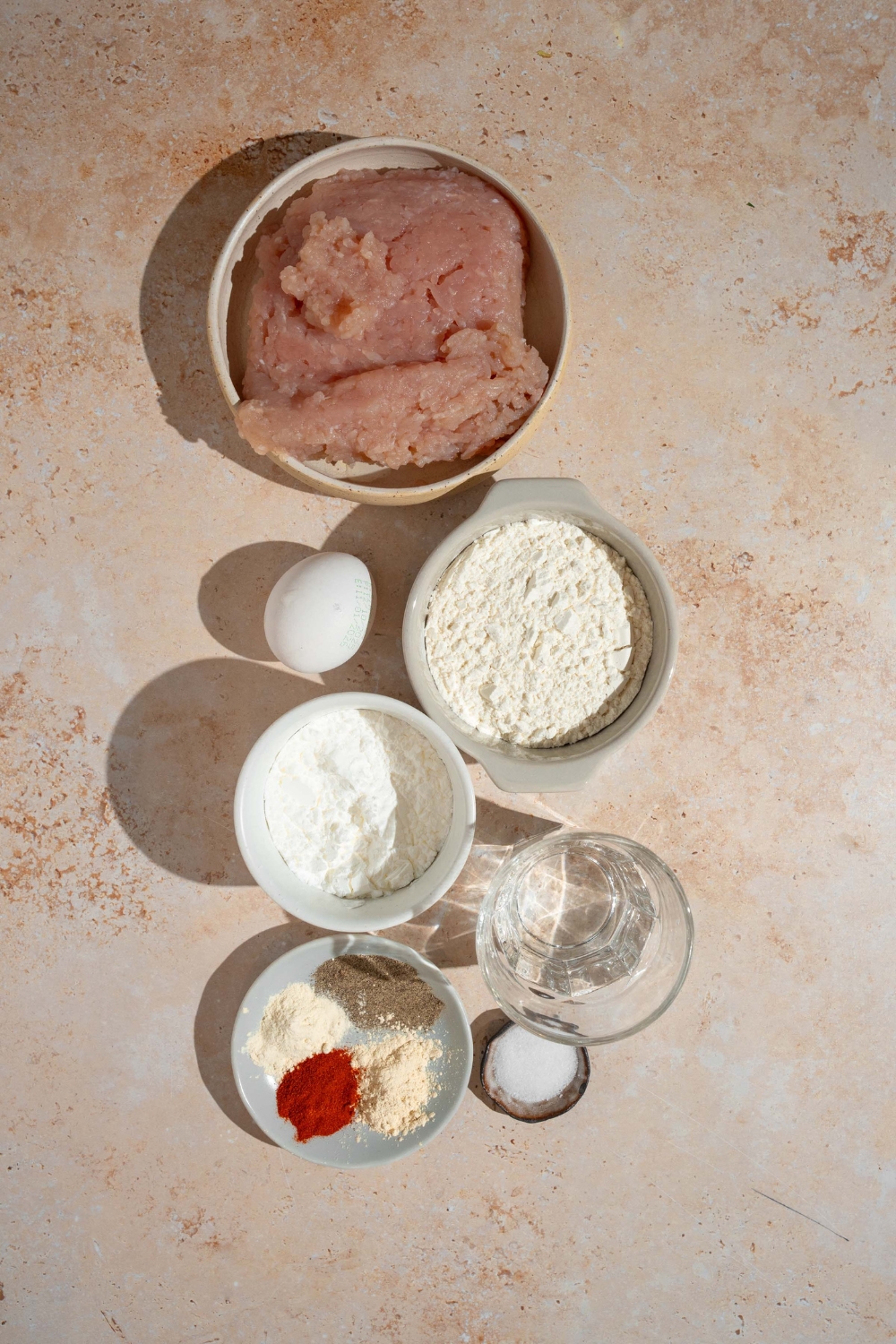 A bowl of ground chicken, a bowl of cornstarch, bowl of flour, egg, a plate with seasonings on it, a bowl of salt, and a bowl of water all on a white counter.
