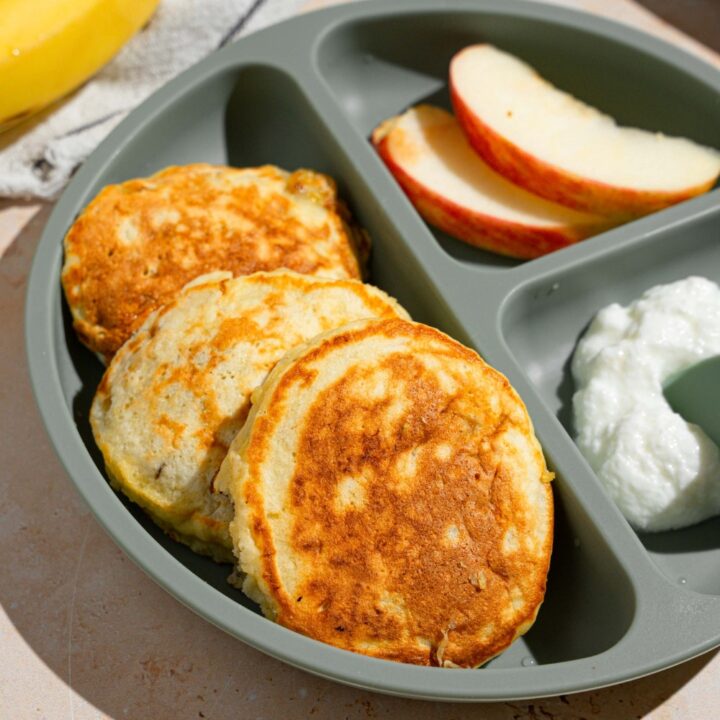 A kids plate with three 3 ingredient banana pancakes with a side of sliced apples and cottage cheese. The plate is on a tan counter with a white striped napkin and bananas.
