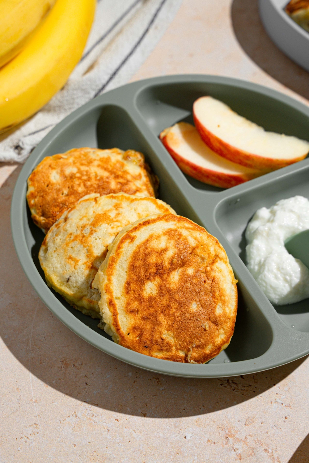 A kids plate with three 3 ingredient banana pancakes with a side of sliced apples and cottage cheese. The plate is on a tan counter with a white striped napkin and bananas.